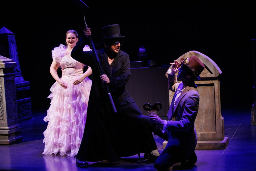 The inventor Coppelius (Kyle Hancock), in a dramatic black top hat and long coat, attacks the inventor Spalanzani (Will Derusha) with a shovel. The clockwork doll Olympia (Holly Howard) looks on in the background with an eerie smile.