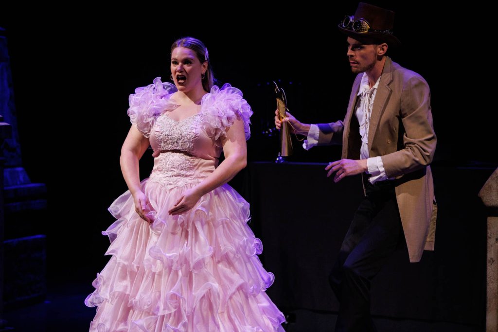 Holly Howard sings the "Doll Aria" from Offenbach's "Les Contes d'Hoffmann." She wears a frilly pink dress, standing in a robotic pose. Will Derusha stands behind her as the inventor Spalanzani, watching her and holding a wind-up attachment.
