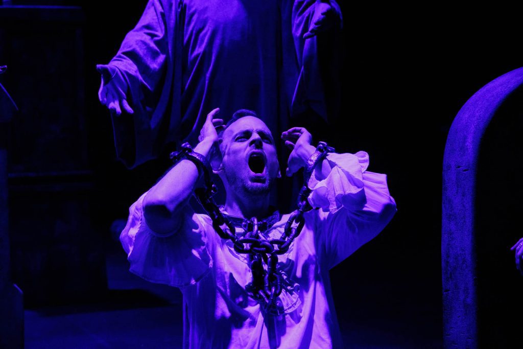 Don Giovanni (Will Derusha), lit in dramatic purple and wearing a white poet's blouse and chains, shouts up to the sky as the Commendatore (Kyle Hancock) looms behind him.