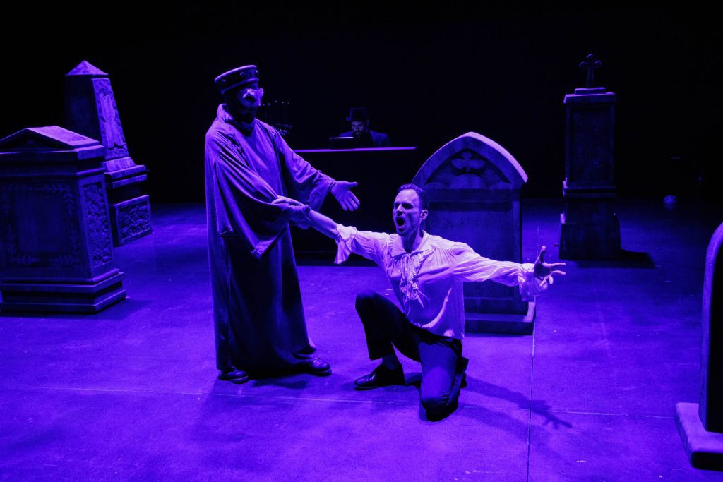 Don Giovanni (Will Derusha) kneels singing before the Commendatore (Kyle Hancock) in dramatic purple lighting, surrounded by tombstones. In the background, Thiago Nascimento accompanies on piano.