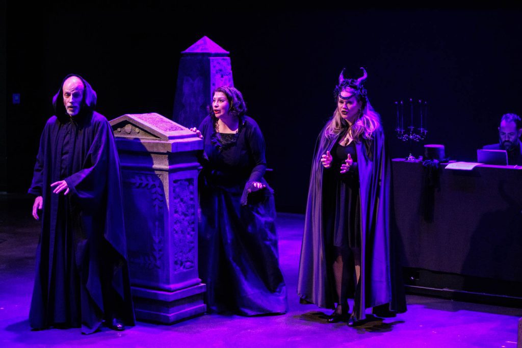 Doug Brunker, Yana White, and Holly Howard perform "Chorus of Witches and Goblins" from "The Vampire," wearing dramatic black robes and standing among tombstones. In the background, with a candelabra on the piano, Thiago Nascimento accompanies.