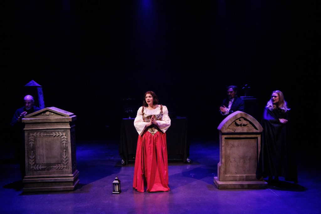 Emmy (Yana White) sings "Emmy’s Ballad of the Vampire" from Heinrich Marschner's "Der Vampyr." She wears a beautiful red peasant gown and a white blouse, looking anxious, with a lantern sitting beside her. Among the tombstones surrounding her, other cast members (Doug Brunker, Kyle Hancock, and Holly Howard) lurk in dark clothing.