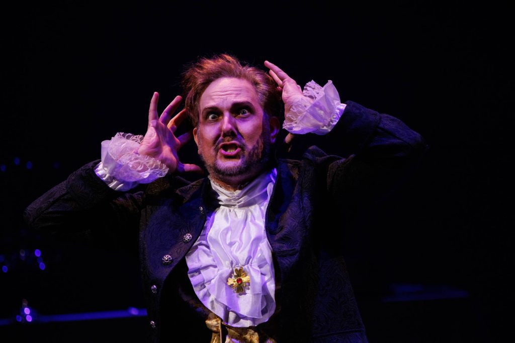 Kyle Hancock, as Sir Roderic Murgatroyd, performs "When the night wind howls" from Gilbert & Sullivan's "Ruddigore." He wears a frilled shirt and a dark jacket, and holds up his hands to his head with a crazed expression.