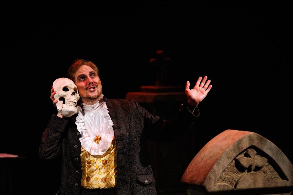 Kyle Hancock, as Sir Roderic Murgatroyd, performs "When the night wind howls" from Gilbert & Sullivan's "Ruddigore." He holds a skull up beside his head and smiles fondly, gesturing outwards with his other hand.