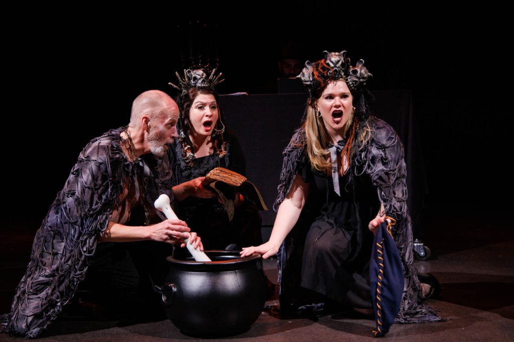 Doug Brunker (right, in a ragged black cape), Yana White (center, crowned with blackened bones), and Holly Howard (right, with skulls in her hair and a similar black cloak), perform the "Witches' Trio" from Verdi's "Macbeth." Doug stirs a black cauldron with a large bone, and Yana reads from a spellbook.