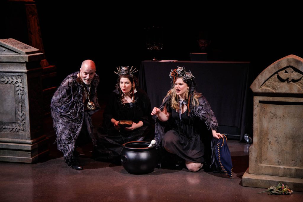 Doug Brunker (right, in a ragged black cape), Yana White (center, crowned with blackened bones), and Holly Howard (right, with skulls in her hair and a similar black cloak), perform the "Witches' Trio" from Verdi's "Macbeth." The three witches look menacingly out at the audience, kneeling around their cauldron among the tombstones. In the shadows behind them, Thiago Nascimento accompanies on piano.