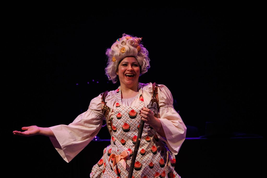 Yana White sings the "Witch's Aria" from Humperdinck's "Hansel and Gretel." She wears a white blouse with a cupcake apron and carries a broom. Her hair is white and fluffy and full of pieces of candy. She smiles broadly at the audience.